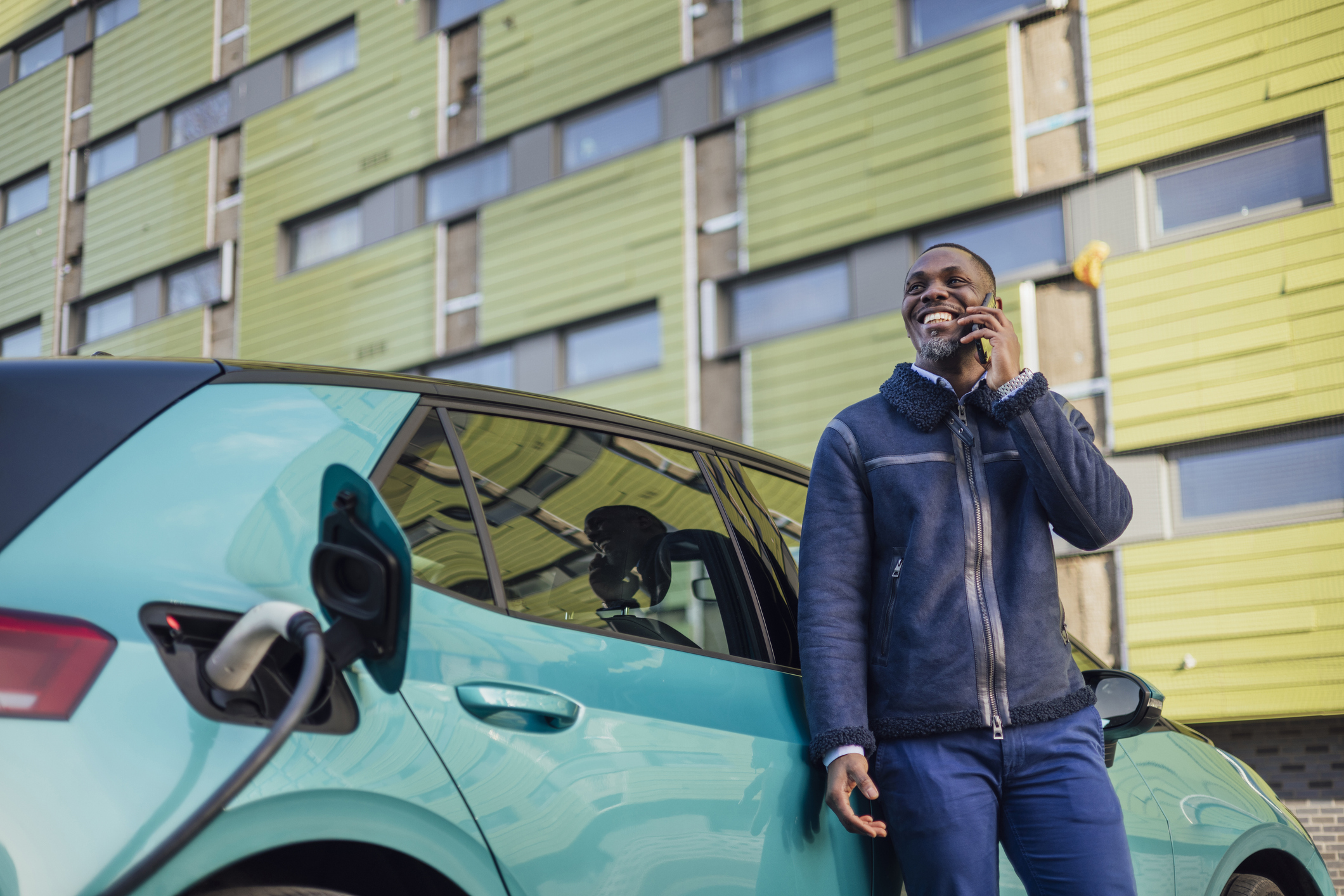  A low angle three quarter length shot of a mid adult businessman wearing formal businesswear taking a phone call on his smartphone while his electric car is on charge at a public charging point in Newcastle upon Tyne in the North East of England.