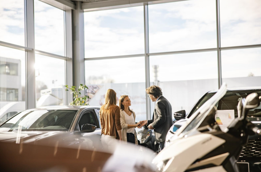 Businessman with short brown hair and glasses is giving the keys to a female customer with long brown hair, both smiling at each other, woman with blond hair standing in the front, indoors at a car dealership in the evening. 