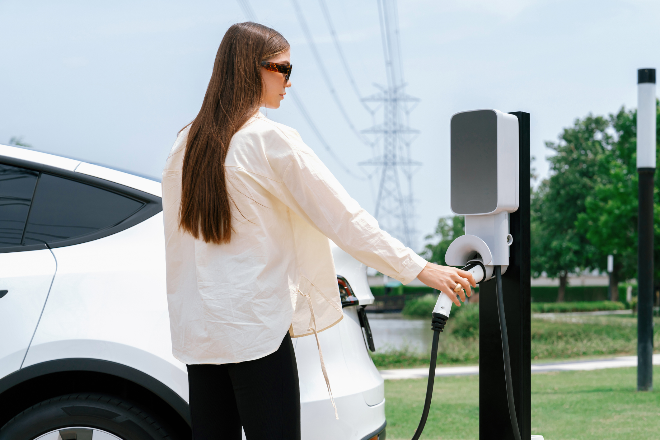 Young woman recharge EV car battery at charging station connected to power grid tower electrical industrial facility as electrical industry for eco friendly vehicle utilisation.