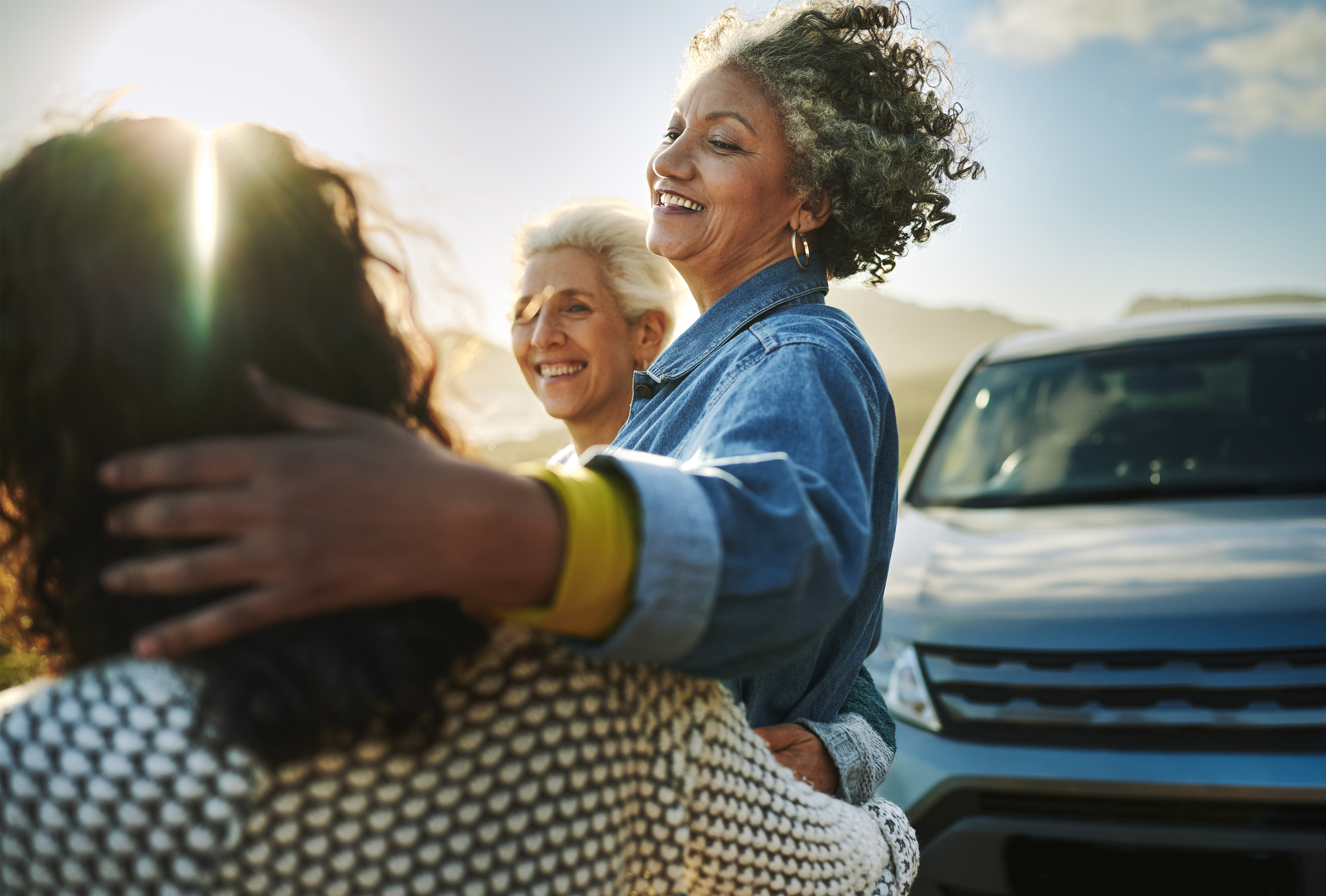 Carefree group of mature female friends standing arm in arm by their car parked on a road running along a scenic coastline