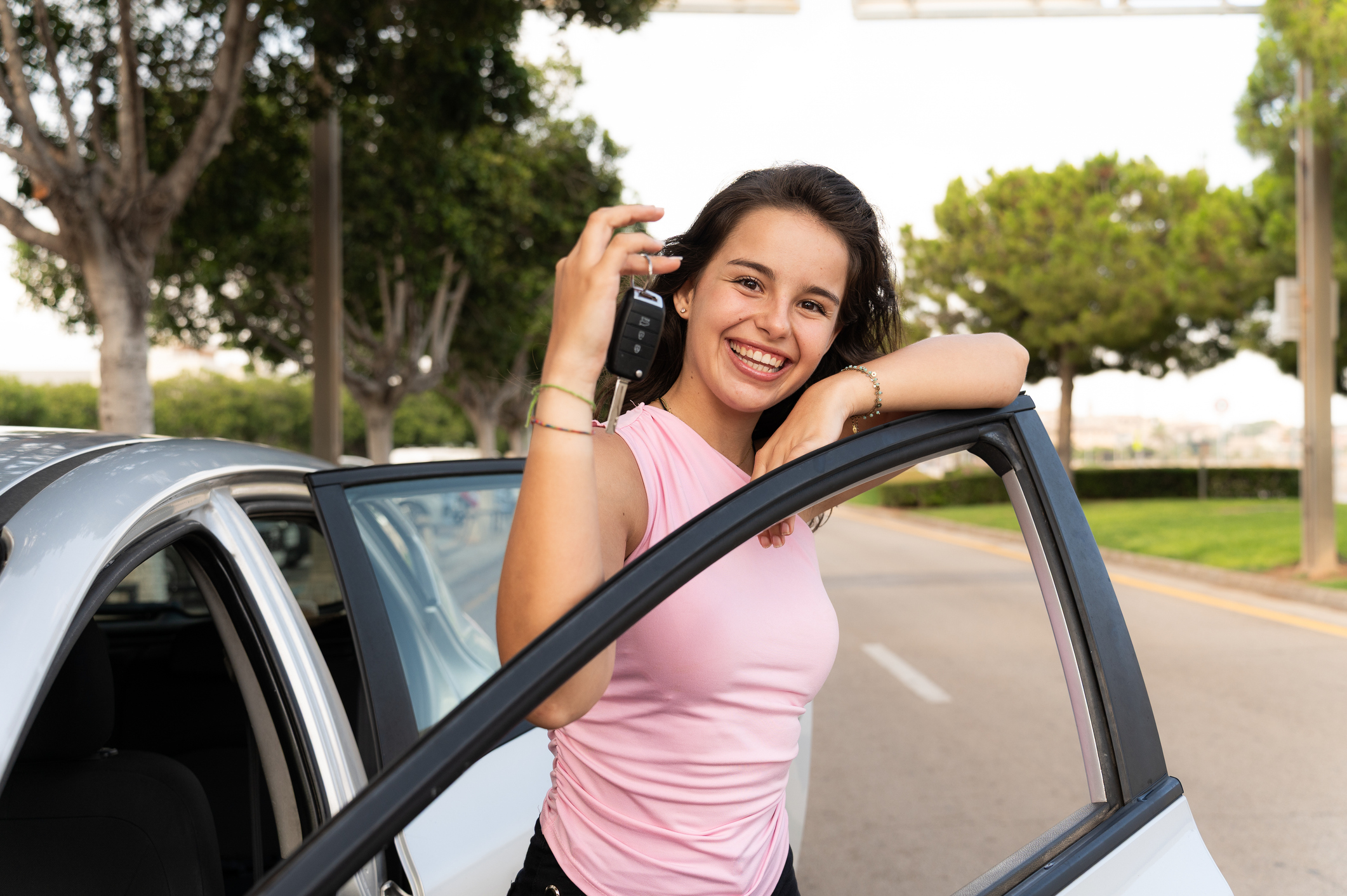 Happy young woman showing car keys while leaning on car door, celebrating purchasing her first vehicle.