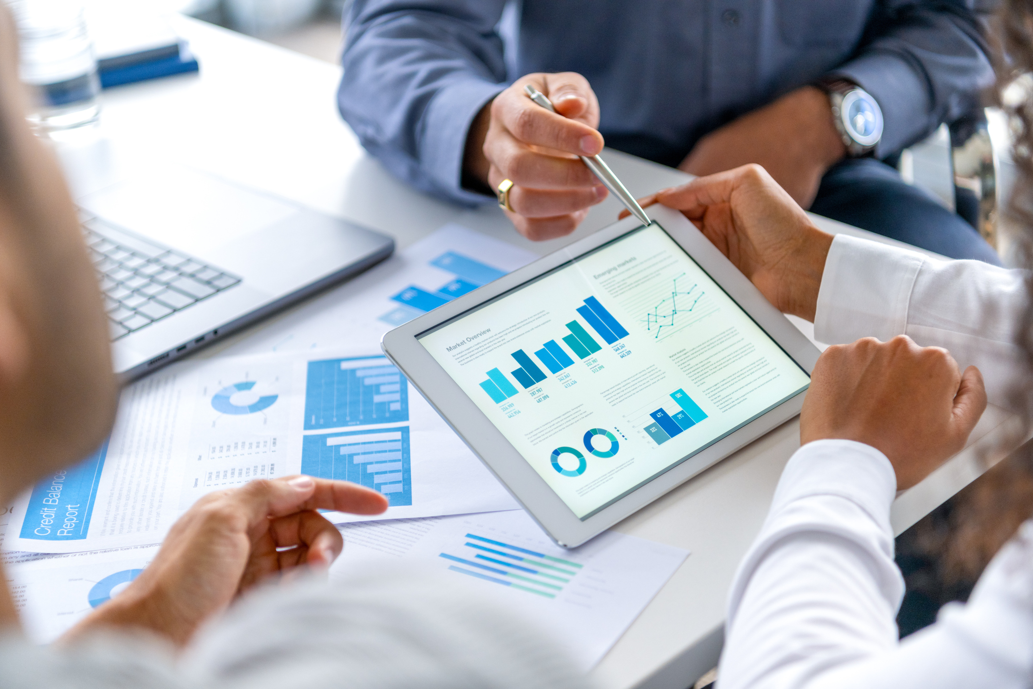 Close up of three people looking at financial data with graphs and charts. All their hands can be seen and one person is pointing with a pen. There is paperwork on the desk showing more finance information.