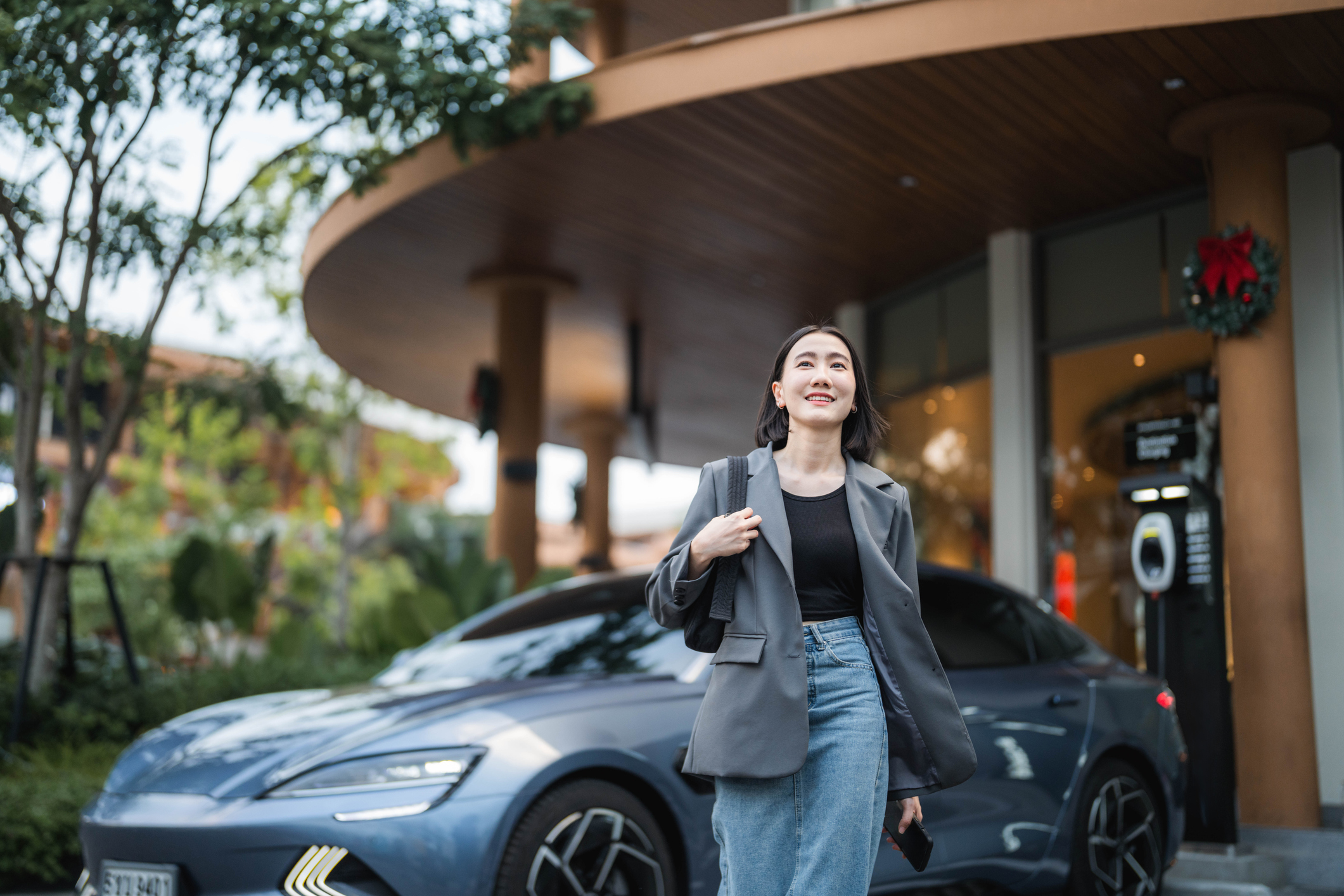 A confident woman stepping out of her EV car, smoothly closing the car door behind her and locking the car through an application on her smartphone. 