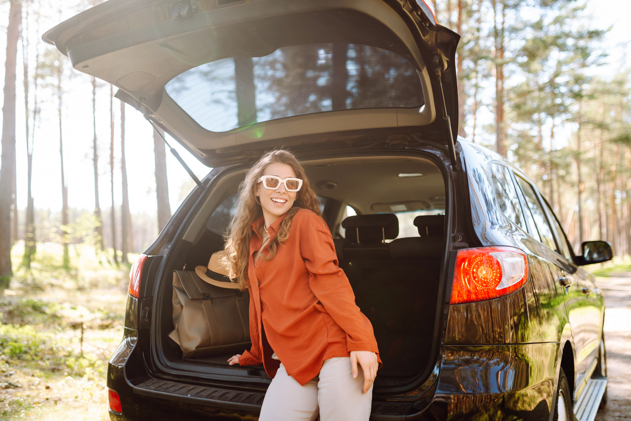  Joyful woman sits in the trunk of a car at sunset in the forest. Traveler enjoys nature, feels freedom. Road trip, weekend concept. Active lifestyle. 