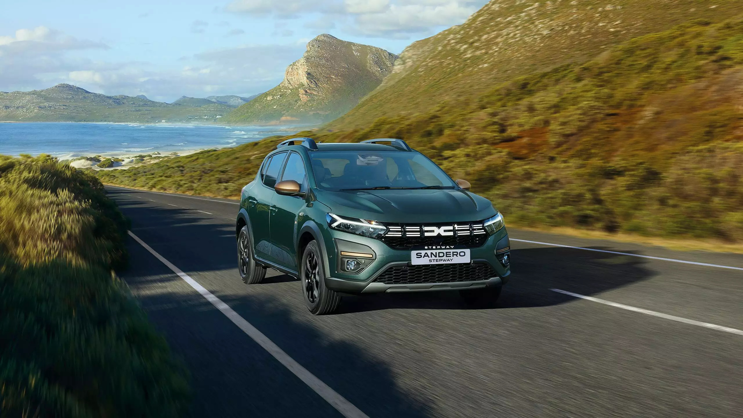 A green Dacia Sandero Stepway hatchback car driving along a rural, mountainous road