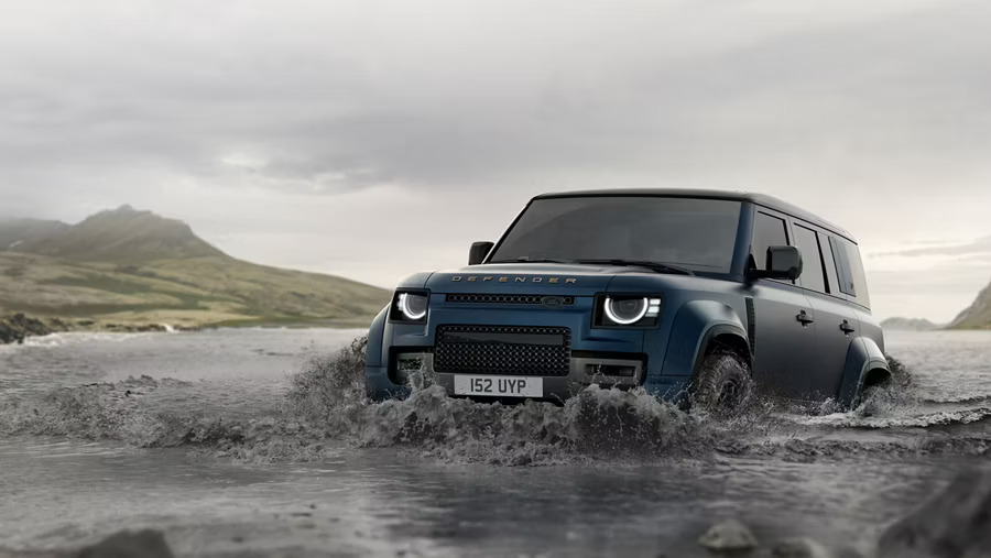 A blue Land Rover Defender driving through deep water with ease. A dark mountainous background.