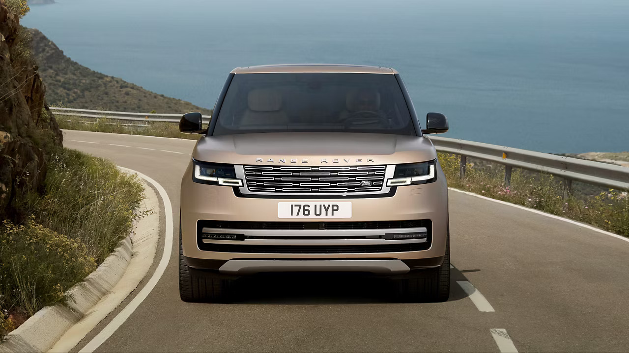 The front view of a gold Range Rover driving along a mountainous road with an ocean view.