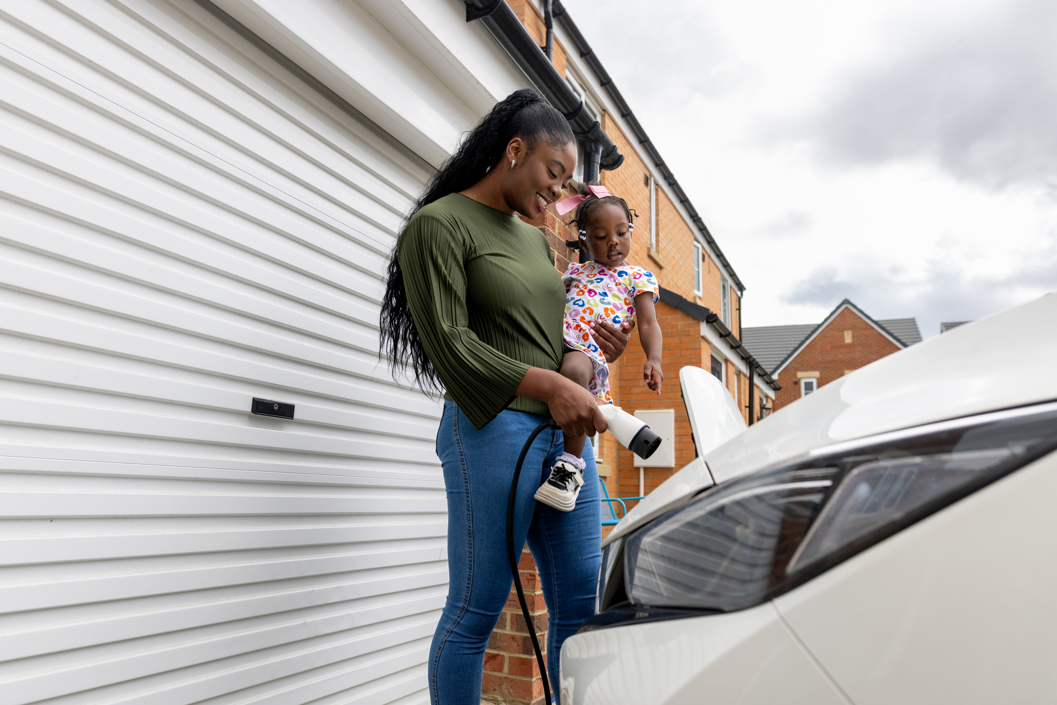 A wide shot of a mother plugging her electric charger into her car while carrying her young daughter in her arms. They are both wearing casual clothing, and the woman is standing in front of her car, which is parked outside her home.