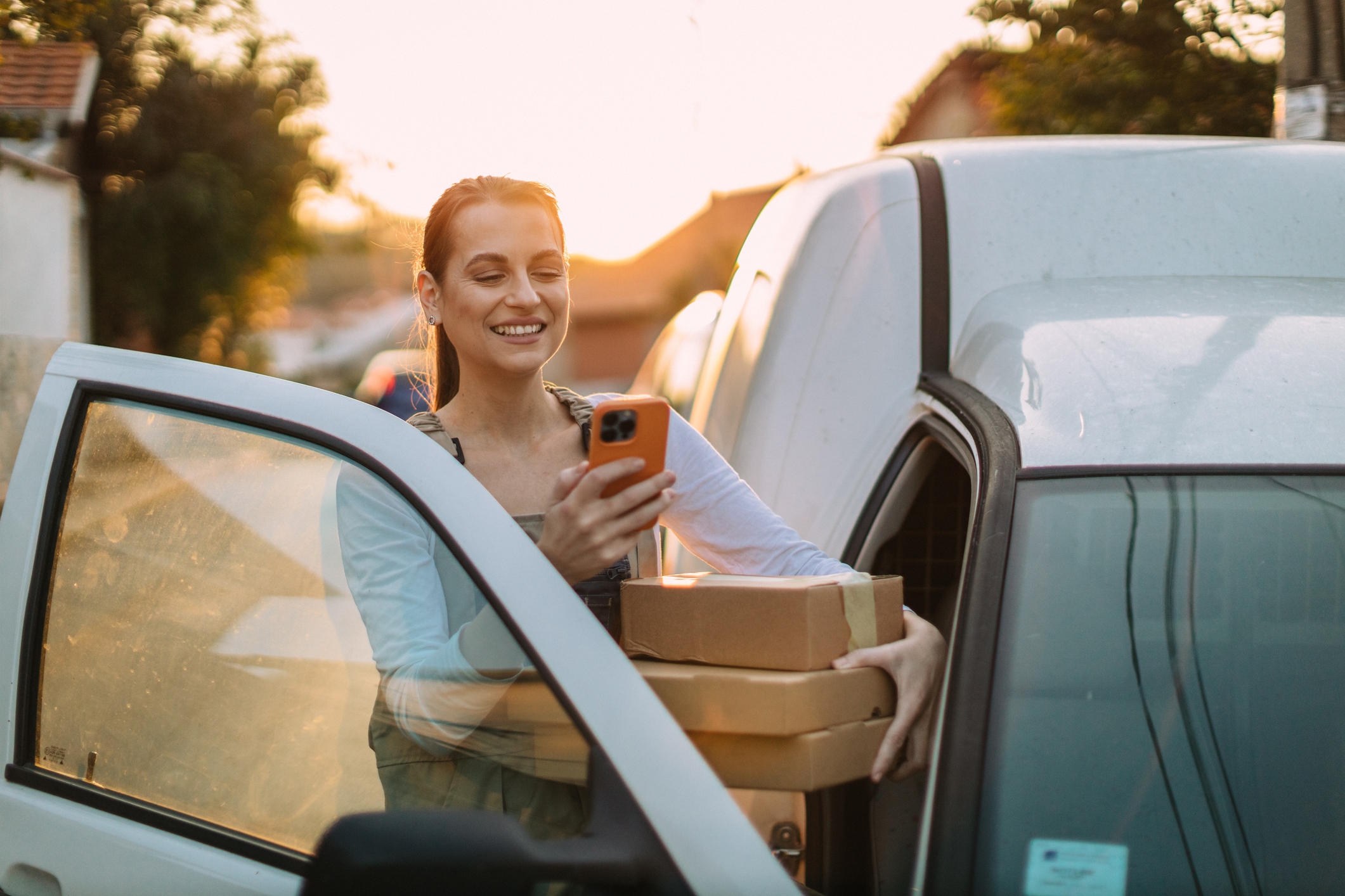 A young woman wearing a uniform using a smartphone and delivering packages in a white van on a sunny day in the city street.