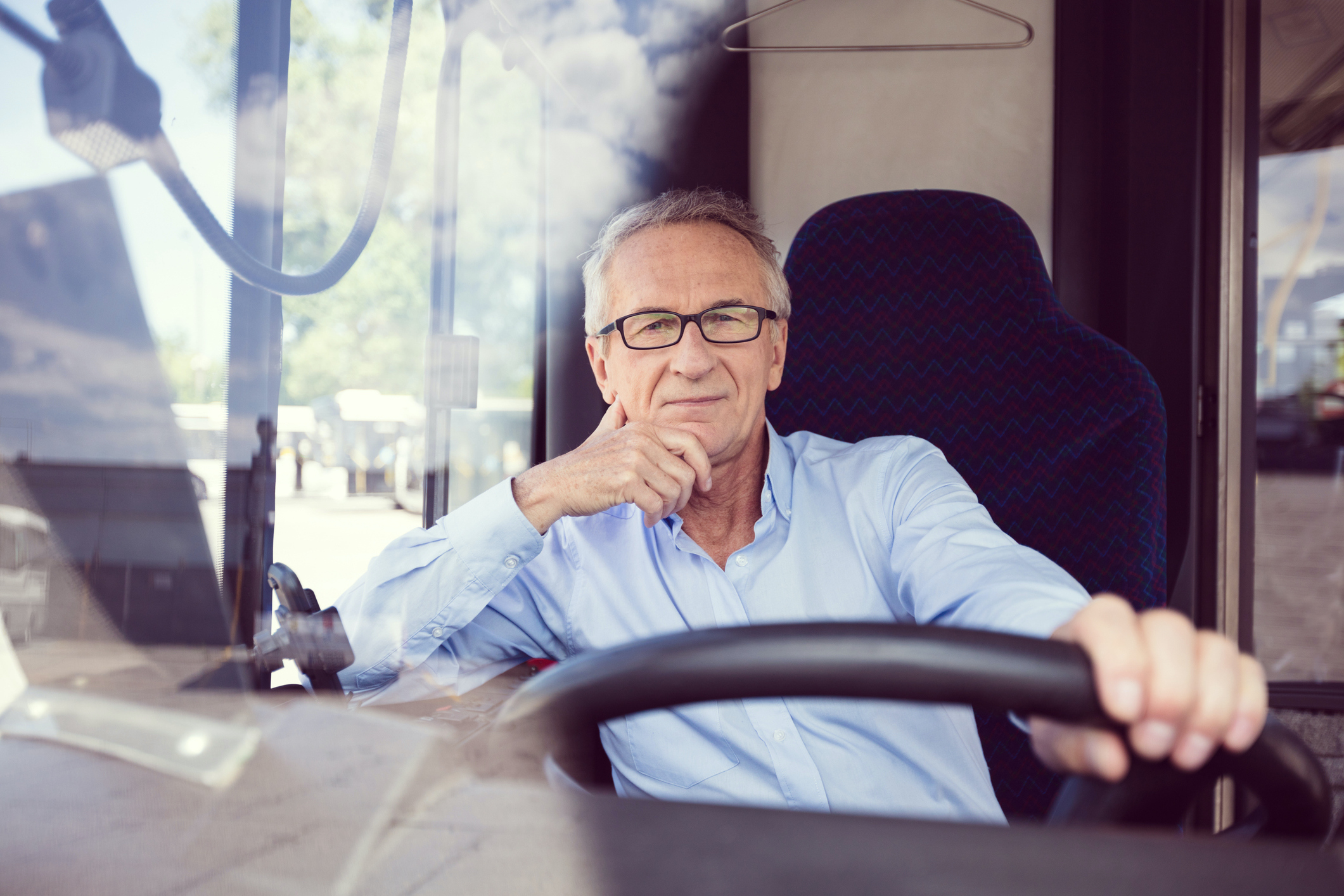Portrait of confident senior man driving bus. Male driver is sitting with hand on chin in public transport. He is wearing formal shirt.