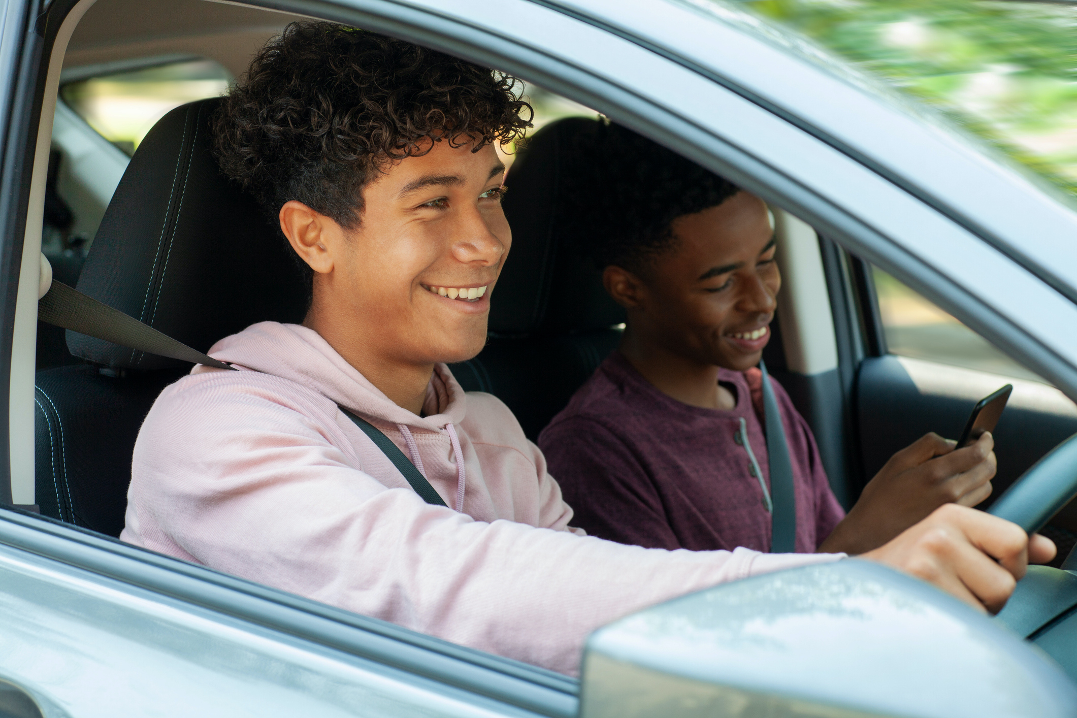 Young Multiracial Hispanic Caucasian teenager boy driving car while Black ethnicity friend uses smart phone while sitting in car with friend passenger wearing seat belt.  