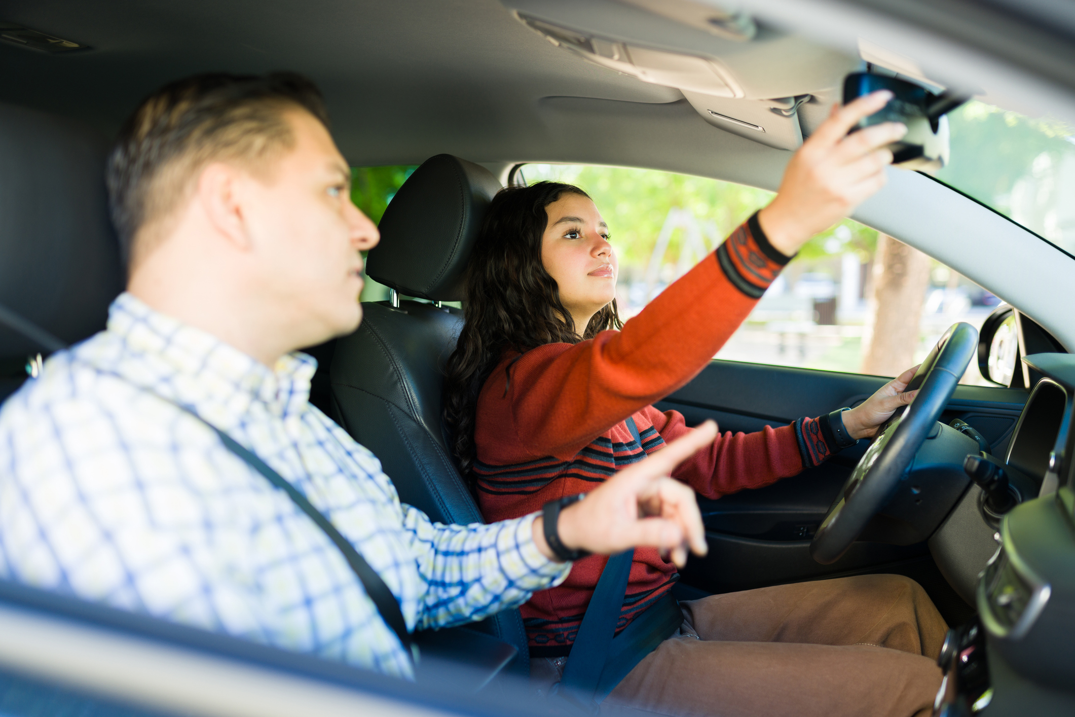 Hispanic teen girl adjusting rearview mirror while learning to drive with her father next to her.